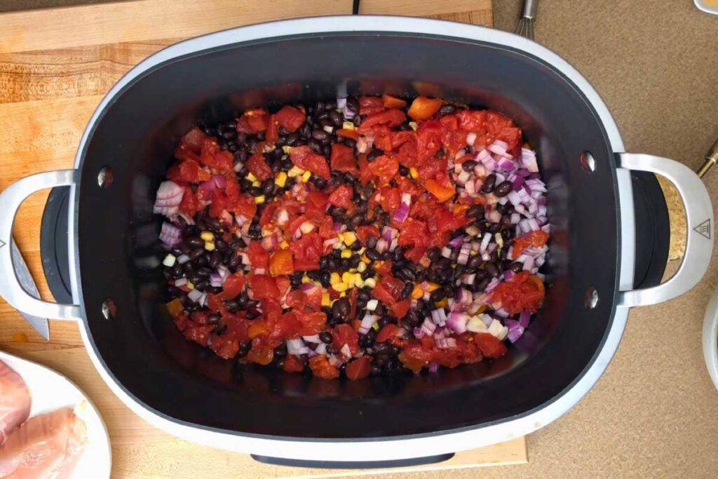 A slow cooker filled with diced tomatoes, black beans, corn, and chopped red onions, prepared for cooking.