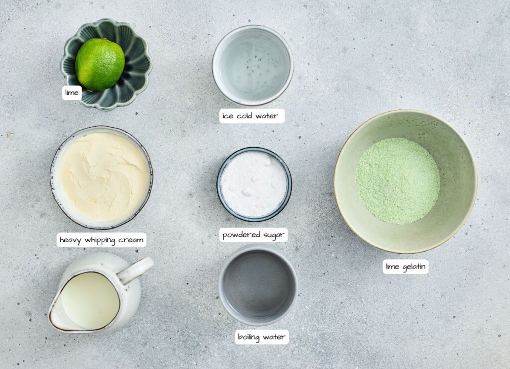 Top-down view of ingredients for a dessert: a whole lime, ice cold water, heavy whipping cream, powdered sugar, lime gelatin powder, boiling water, and milk in separate bowls.