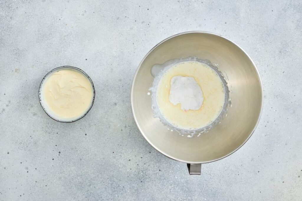 A metal mixing bowl containing cream and a white substance next to a smaller bowl of softened cream cheese on a light gray textured surface.
