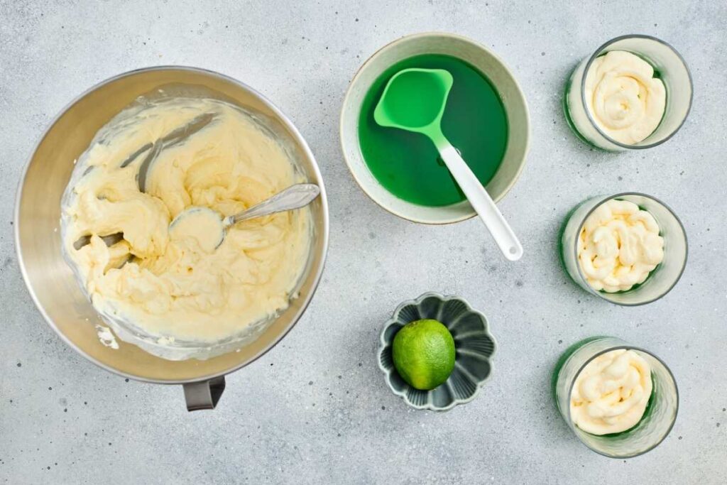 A mixing bowl with creamy batter, a bowl of green liquid with a spatula, a lime in a dish, and three glasses of mousse on a light surface.