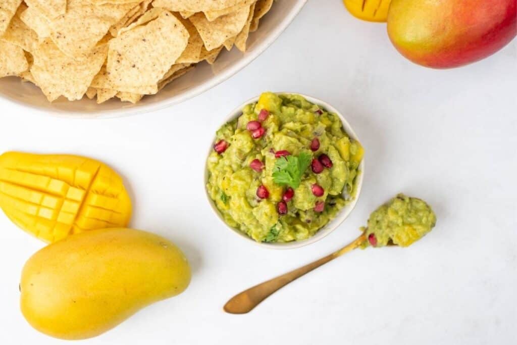 A bowl of Tropical mango guacamole sits next to a spoon, tortilla chips, and two mangoes on a white surface.