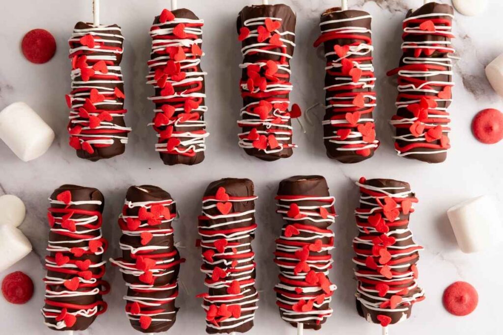 Ten chocolate-covered marshmallow sticks topped with red heart-shaped sprinkles and white drizzle, arranged on a white surface with loose marshmallows and candies around them.