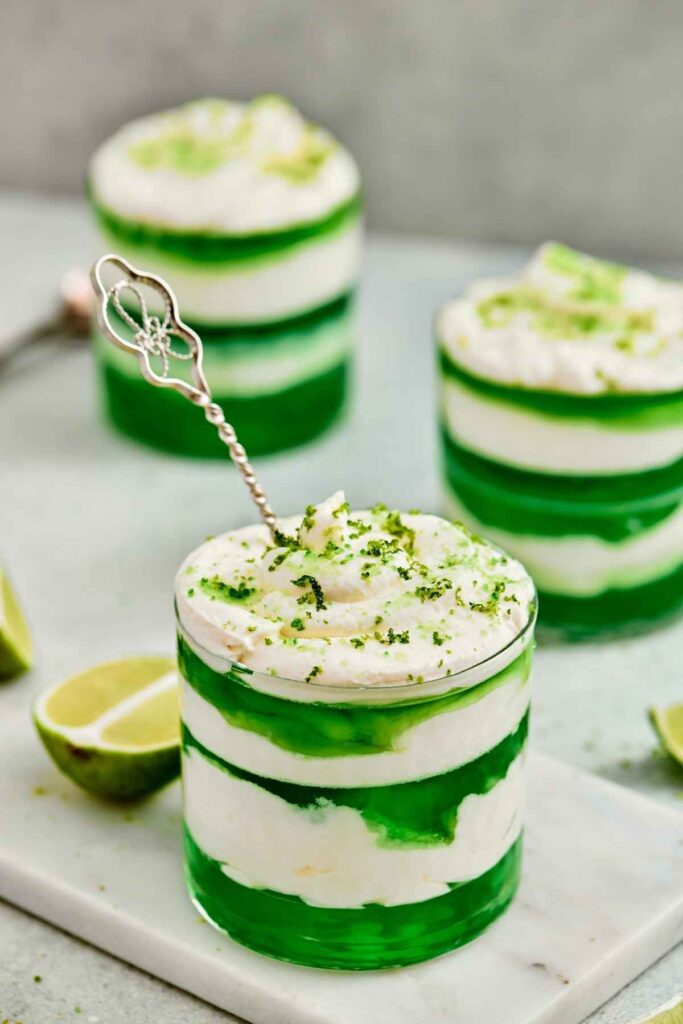 A glass dessert with alternating green jelly and white cream layers, topped with whipped cream and lime zest, with slices of lime nearby.