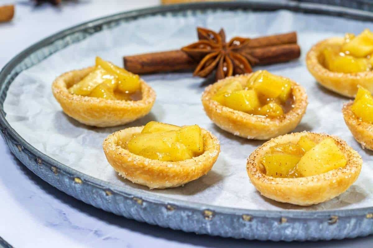 Mini apple tartlets with diced apple filling are arranged on a round metal tray, garnished with a cinnamon stick and a star anise in the background.