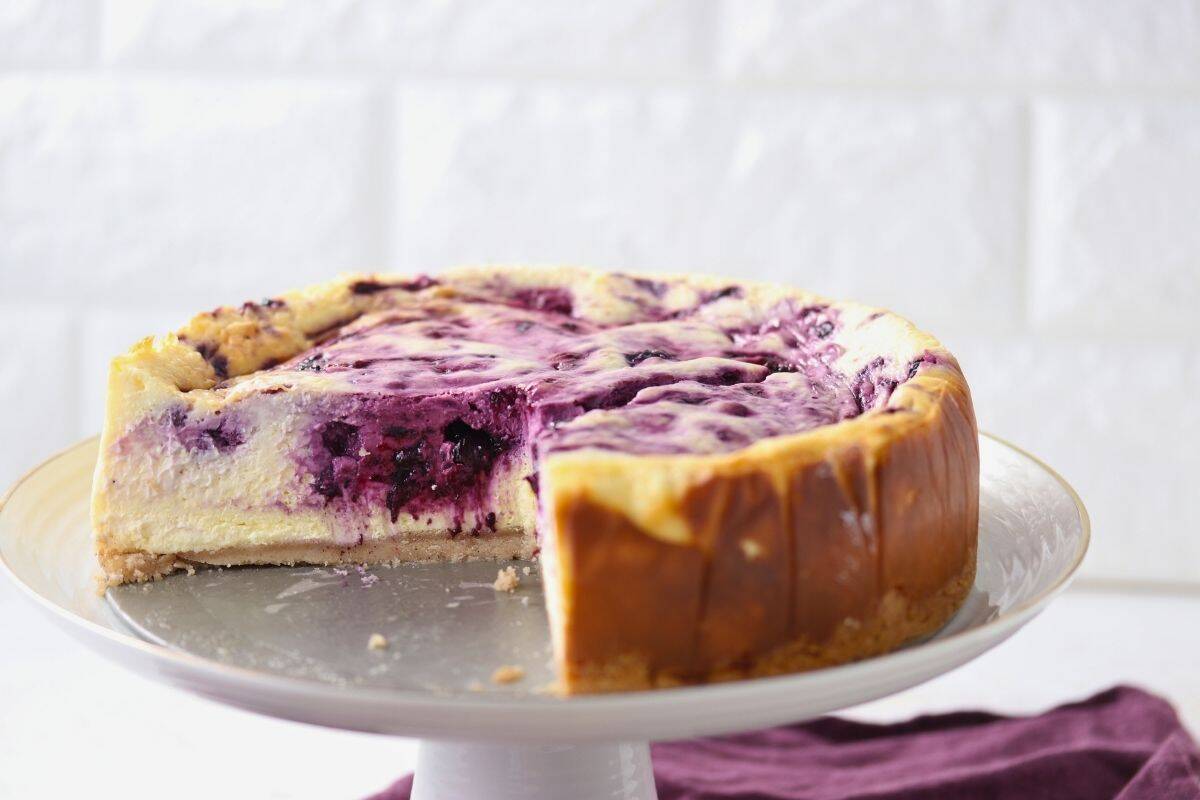 A blueberry swirl cheesecake with a slice removed, displayed on a white cake stand against a light background.