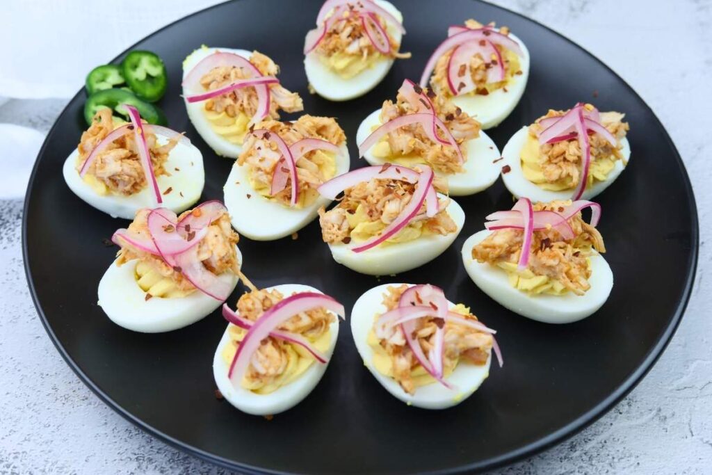 A black plate with twelve deviled eggs topped with shredded meat and pickled red onions, garnished with sliced jalapeños on the side.