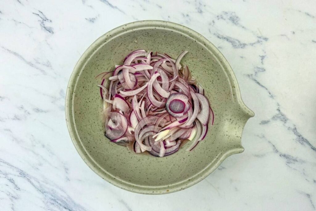 A speckled mixing bowl containing thinly sliced red onions sits on a white marble surface.