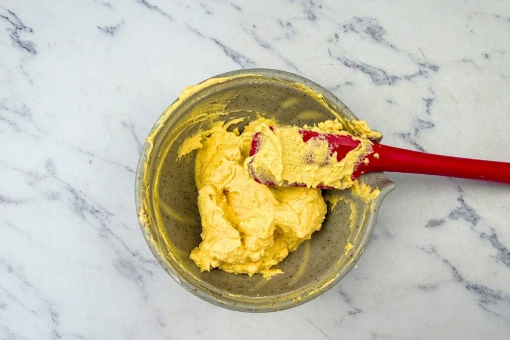 A mixing bowl with yellow dough and a red spatula on a white marble countertop.