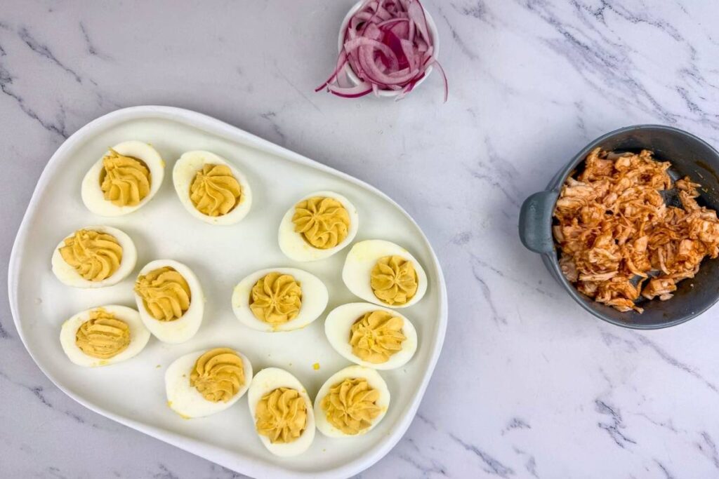 A plate of deviled eggs, a bowl of shredded chicken, and a small dish of sliced red onions on a white marble surface.