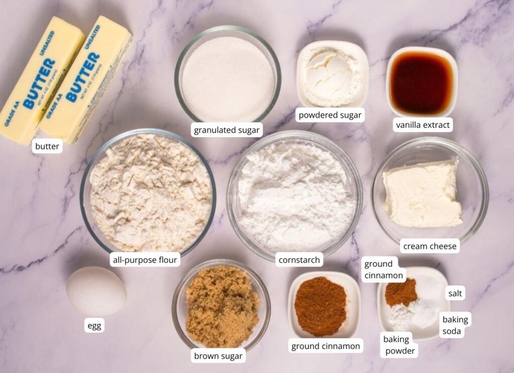 Various baking ingredients labeled in bowls and containers, including butter, sugars, flour, cornstarch, spices, baking powder, baking soda, cream cheese, vanilla extract, and an egg.