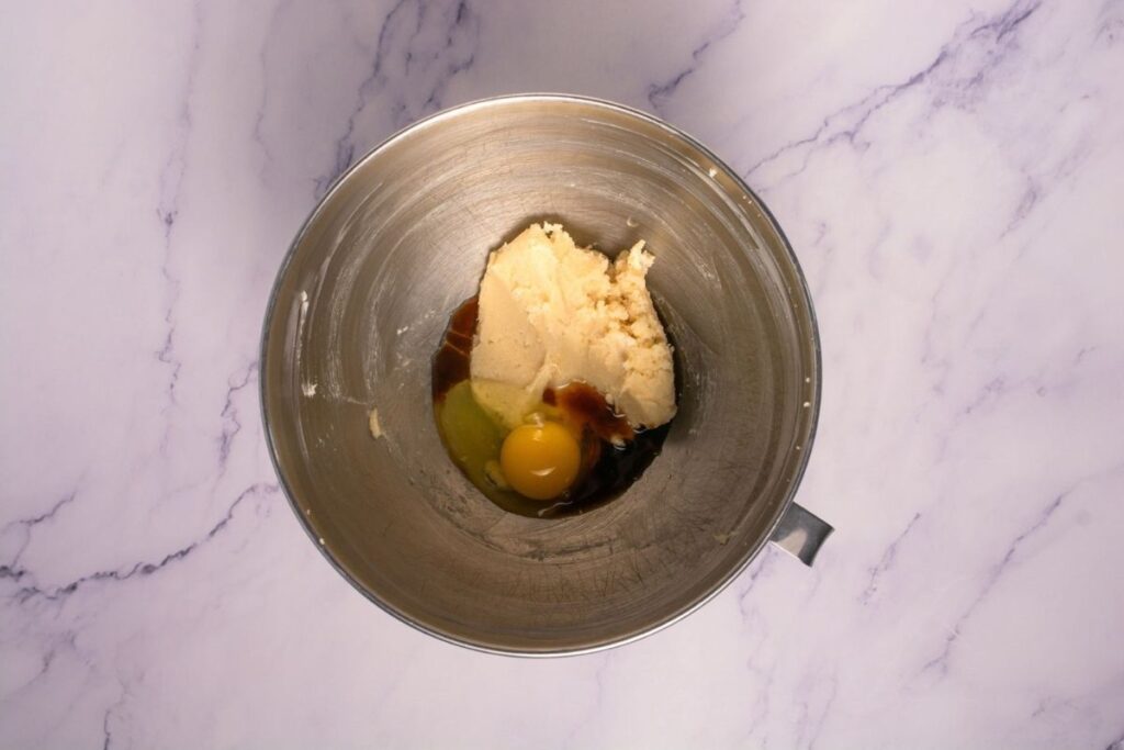 A metal mixing bowl containing butter, sugar, vanilla extract, and a raw egg on a marble countertop.