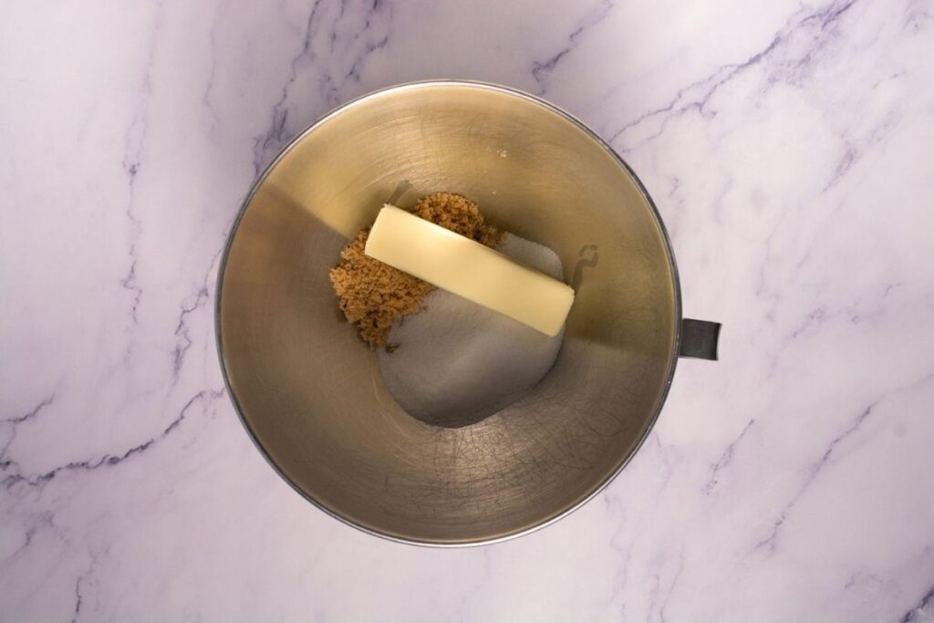 A metal mixing bowl contains a stick of butter, brown sugar, and white sugar on a marble countertop.