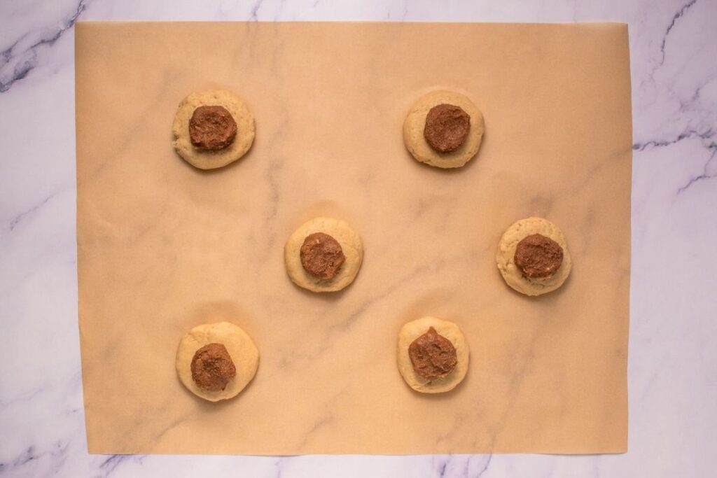 Six unbaked cookies with chocolate centers are arranged on a sheet of parchment paper on a marble surface.