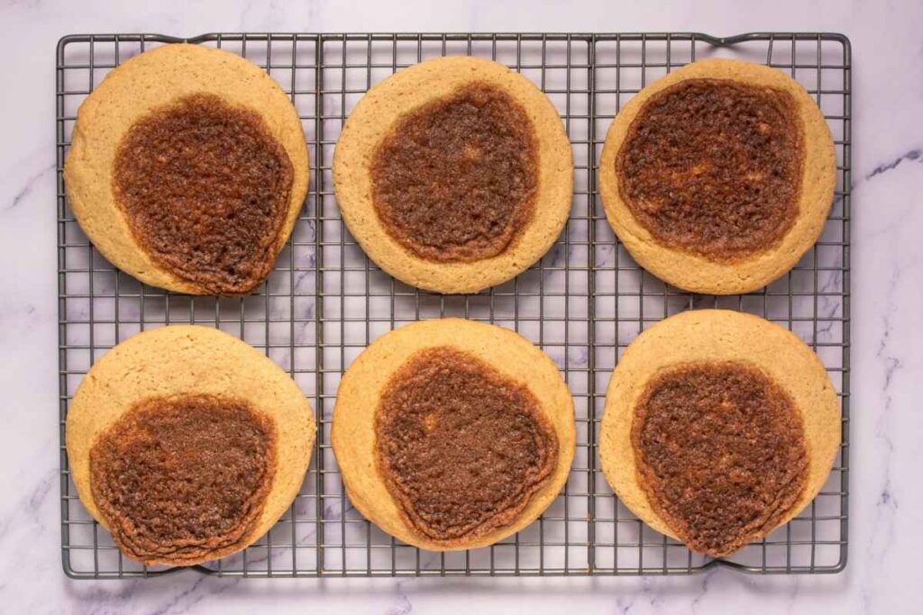 Six large cookies with browned centers are cooling on a wire rack set on a marble surface.