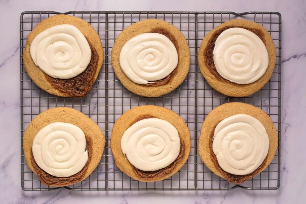 Six large cookies with white icing swirled on top are arranged on a cooling rack over a marble surface.