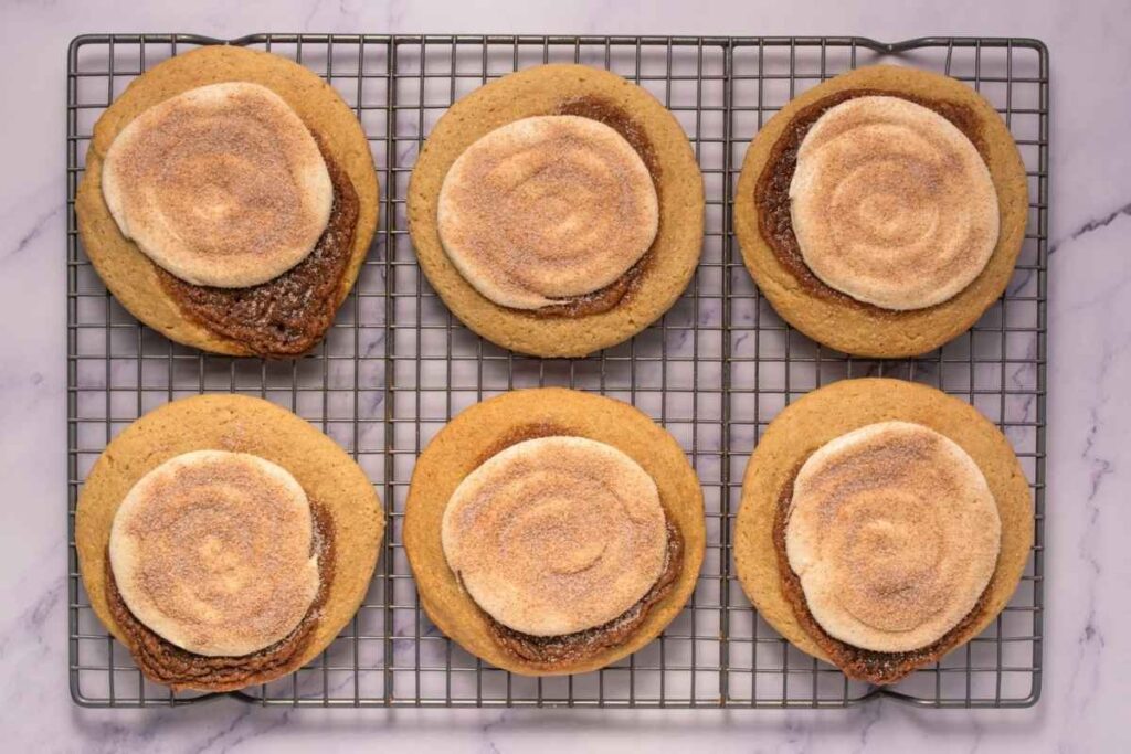 Six round cookies topped with swirled cinnamon and sugar sit on a cooling rack placed on a marble surface.