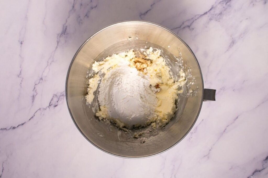 A metal mixing bowl contains creamed butter, sugar, vanilla extract, and flour on a white marble surface.