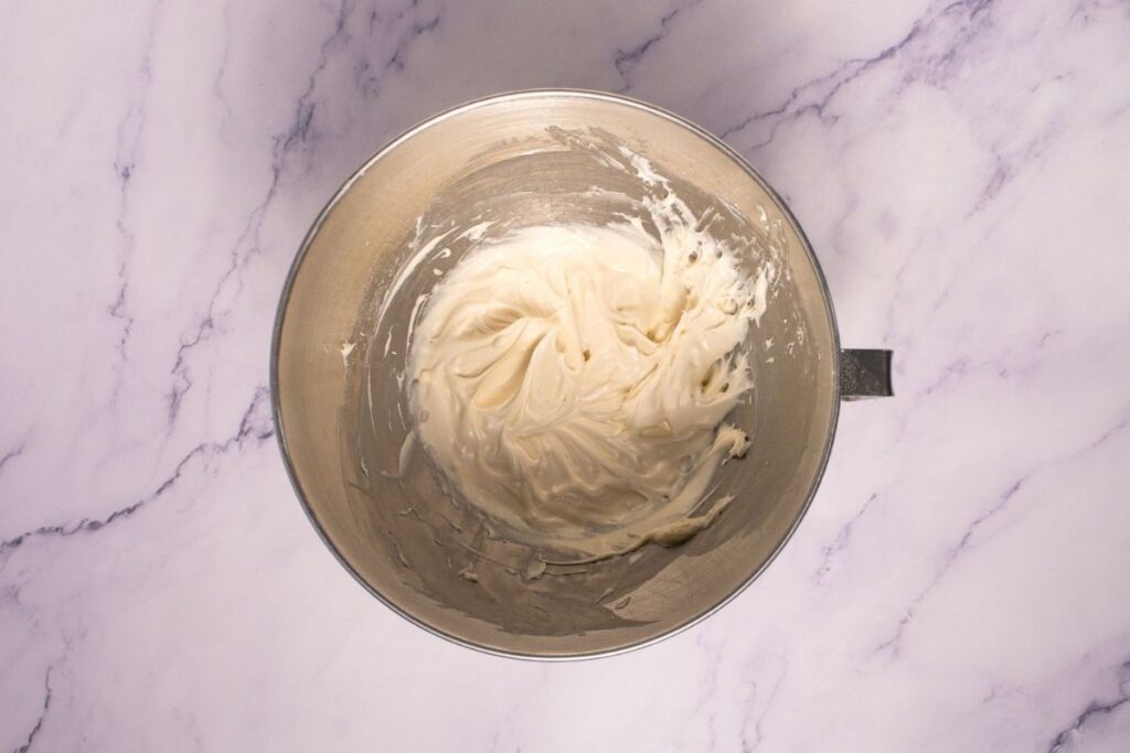A metal mixing bowl containing whipped cream cheese frosting sits on a light marbled surface.