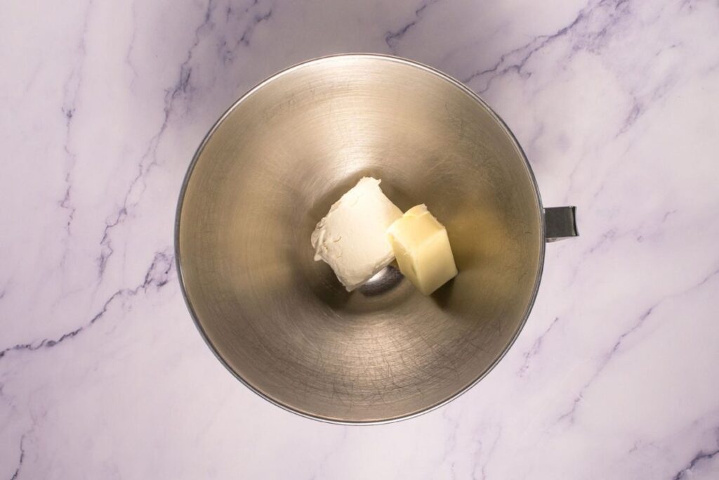 A metal mixing bowl containing a block of cream cheese and a stick of butter, placed on a white marble surface.