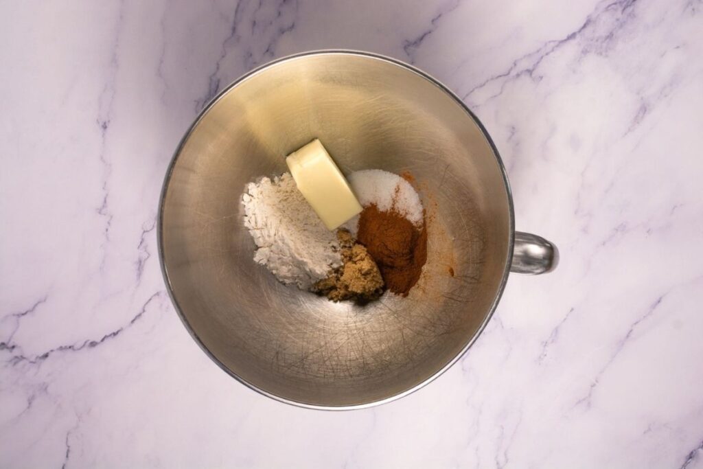A metal mixing bowl with flour, brown sugar, white sugar, cinnamon, and a stick of butter on a white marble surface.