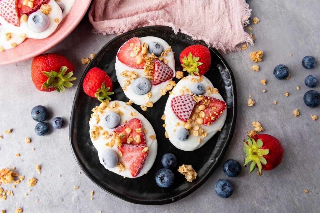 A black plate with yogurt bark pieces topped with sliced strawberries, blueberries, and granola, surrounded by whole strawberries and blueberries on a gray surface.