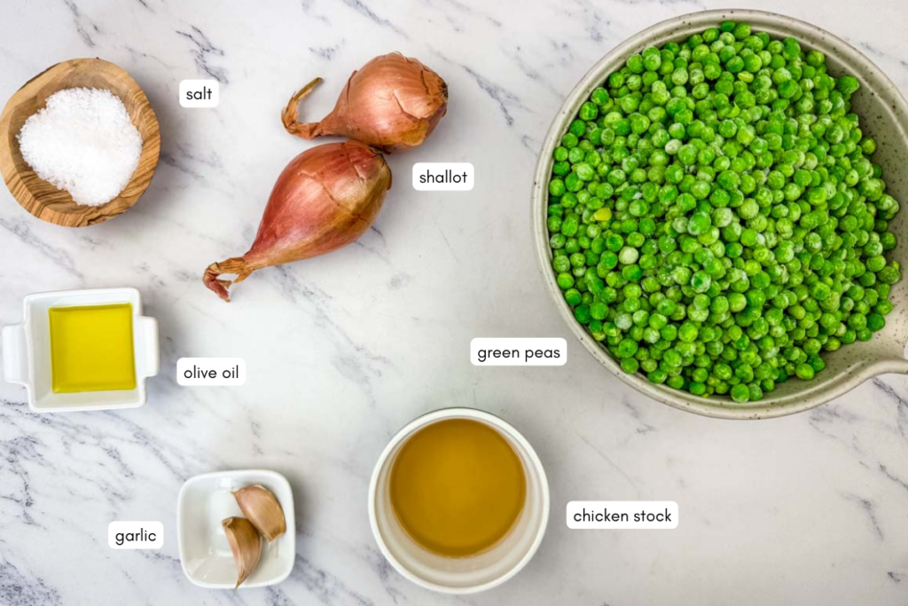 Ingredients for a recipe arranged on a white marble surface: salt, shallot, green peas, chicken stock, garlic, and olive oil.