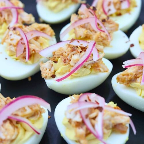 Deviled eggs topped with shredded chicken, red onion slices, and seasoning, arranged on a dark serving platter.