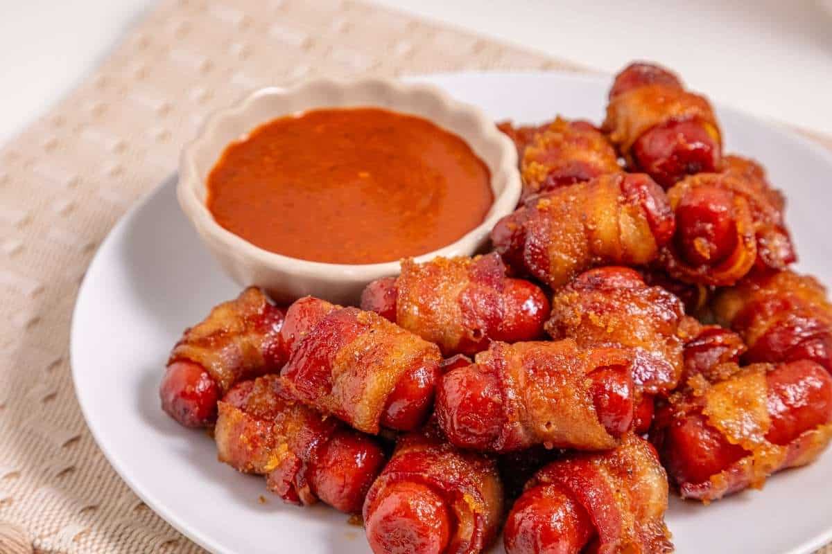 A plate of bacon-wrapped smokies sits next to a small bowl of reddish dipping sauce on a light-colored surface.