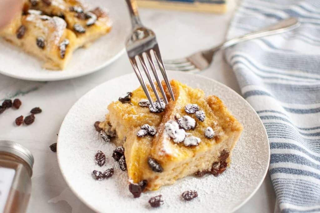 A fork cutting into a slice of bread pudding with raisins, dusted with powdered sugar, served on a white plate.