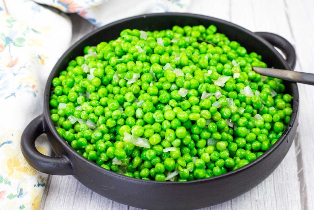 A black bowl filled with cooked green peas and diced onions, with a spoon resting inside, placed on a light wooden surface.
