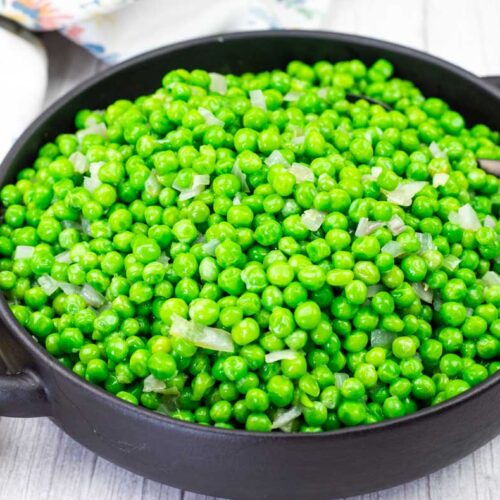 A black bowl filled with cooked green peas and diced onions, with a spoon resting inside, placed on a light wooden surface.