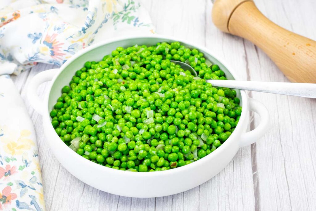 A white bowl filled with cooked green peas and diced onions, with a spoon inside. A floral cloth and a wooden pepper grinder are beside the bowl on a white wooden surface.