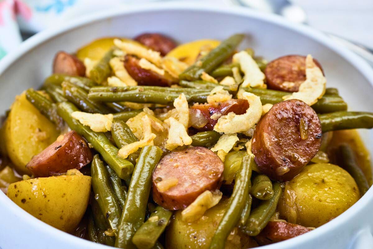A bowl of Swamp Potatoes with cooked green beans, sliced sausage, and halved potatoes, topped with crispy fried onions.