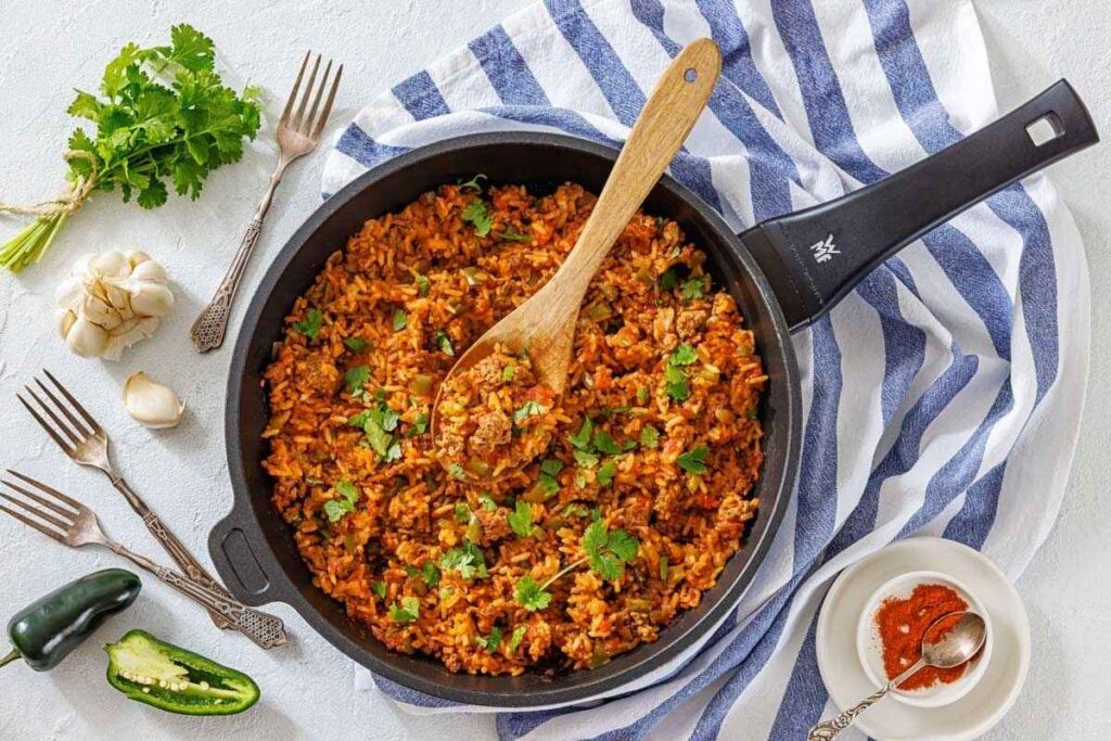 A skillet filled with seasoned rice and ground meat, garnished with cilantro, sits on a striped towel with a wooden spoon. Surrounding it are forks, garlic, jalape&ntilde;o, and spices.