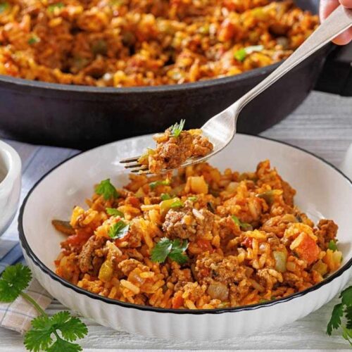 A bowl of rice mixed with ground meat and vegetables is garnished with cilantro; a fork lifts a portion, and a skillet with more of the dish is in the background.