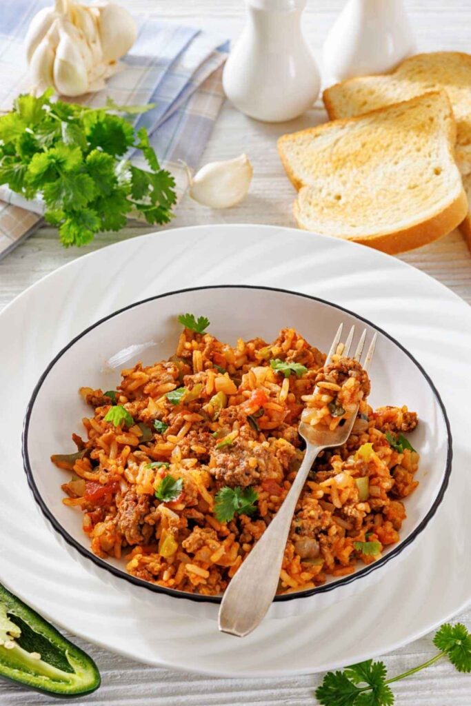 A plate of seasoned rice with ground meat, vegetables, and cilantro, served with toasted bread; a fork rests in the rice. Garlic, parsley, and pepper are arranged around the plate.