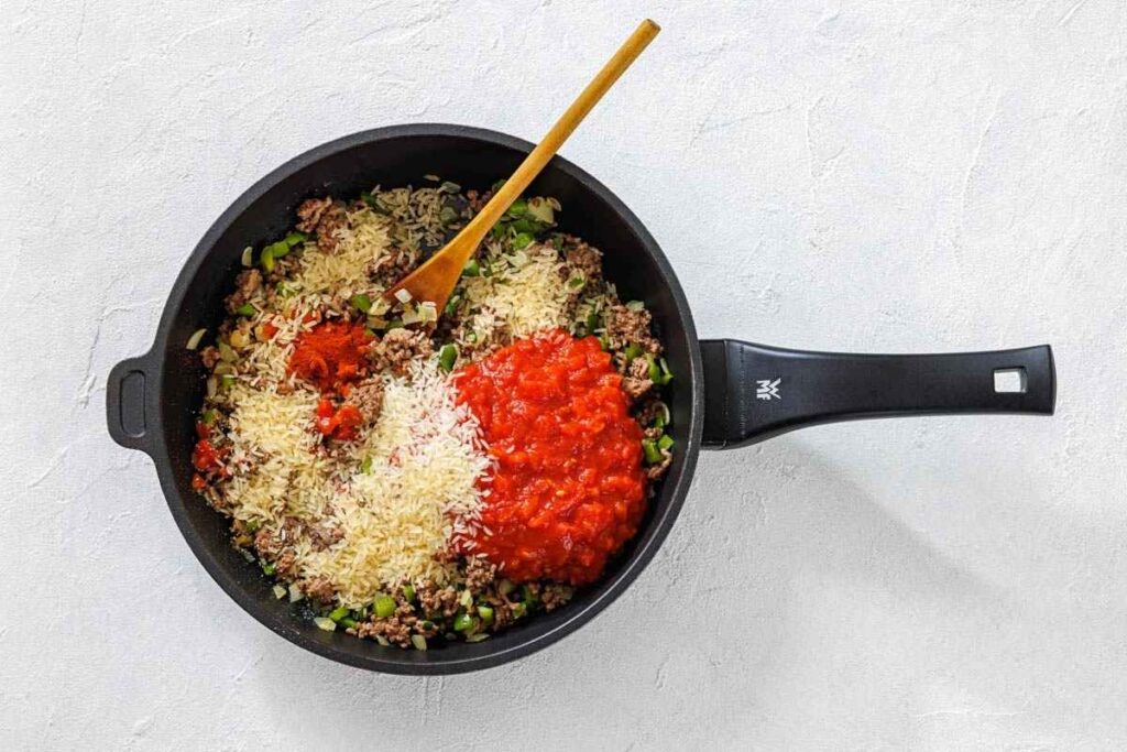 A black skillet contains ground meat, chopped green peppers, uncooked rice, crushed tomatoes, and spices, being mixed with a wooden spoon on a white surface.
