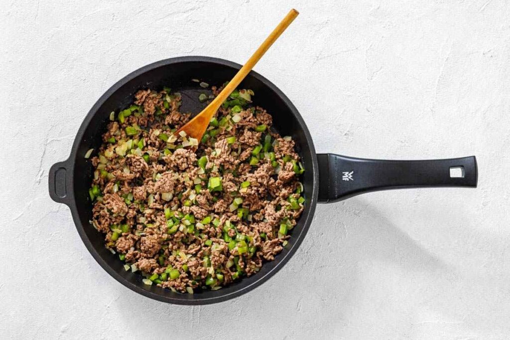 A black skillet containing cooked ground beef mixed with chopped green bell peppers and onions, with a wooden spoon resting inside.