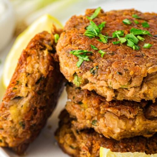 Three golden-brown patties stacked on a plate, garnished with chopped herbs, with lemon wedges and a bowl of white sauce in the background.