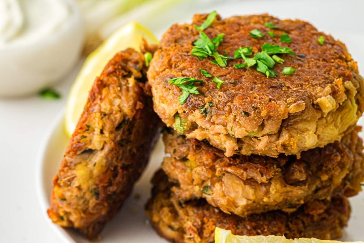 Three golden-brown patties stacked on a plate, garnished with chopped herbs, with lemon wedges and a bowl of white sauce in the background.