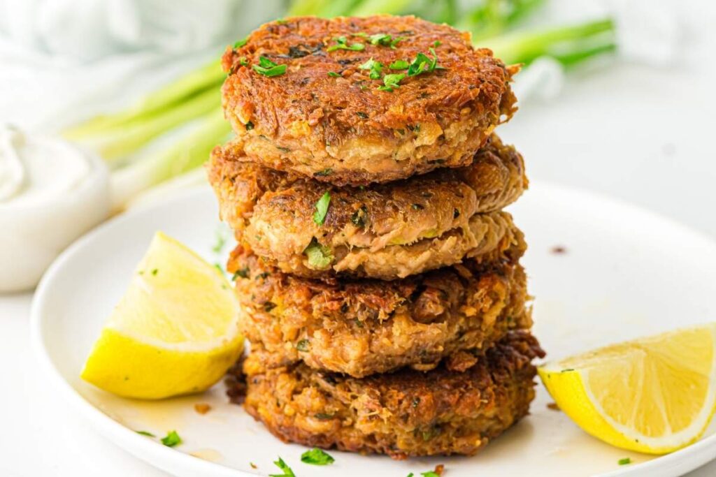A stack of four golden-brown patties garnished with herbs on a white plate, served with lemon wedges.