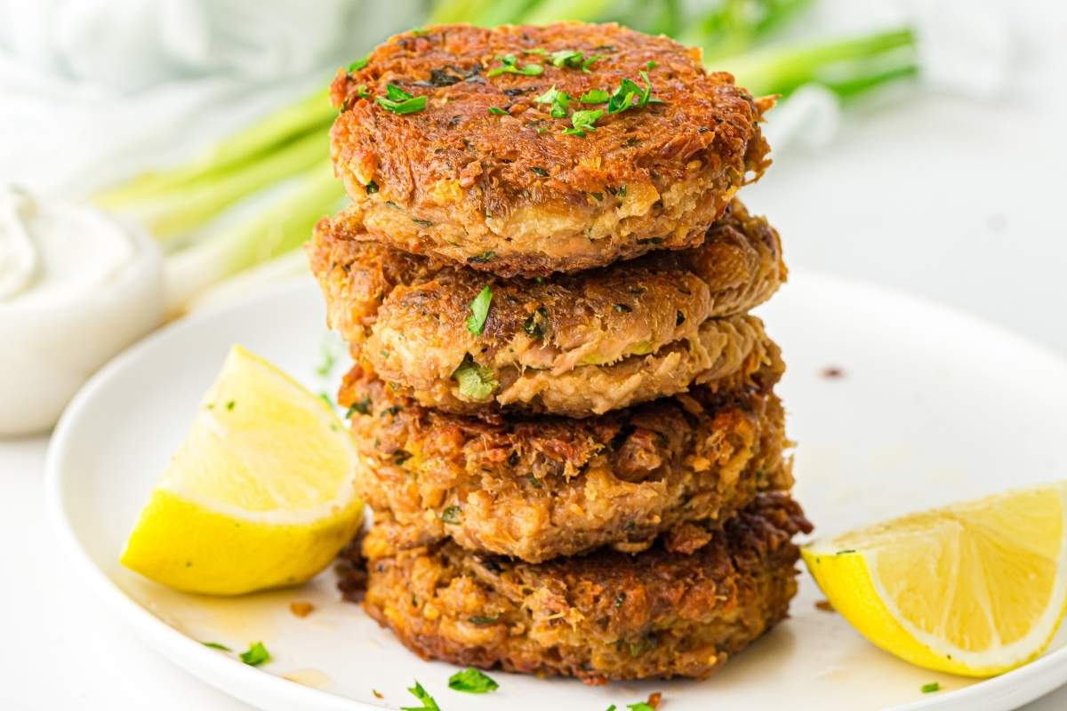 A stack of four golden-brown tuna patties garnished with chopped herbs, served on a white plate with two lemon wedges.