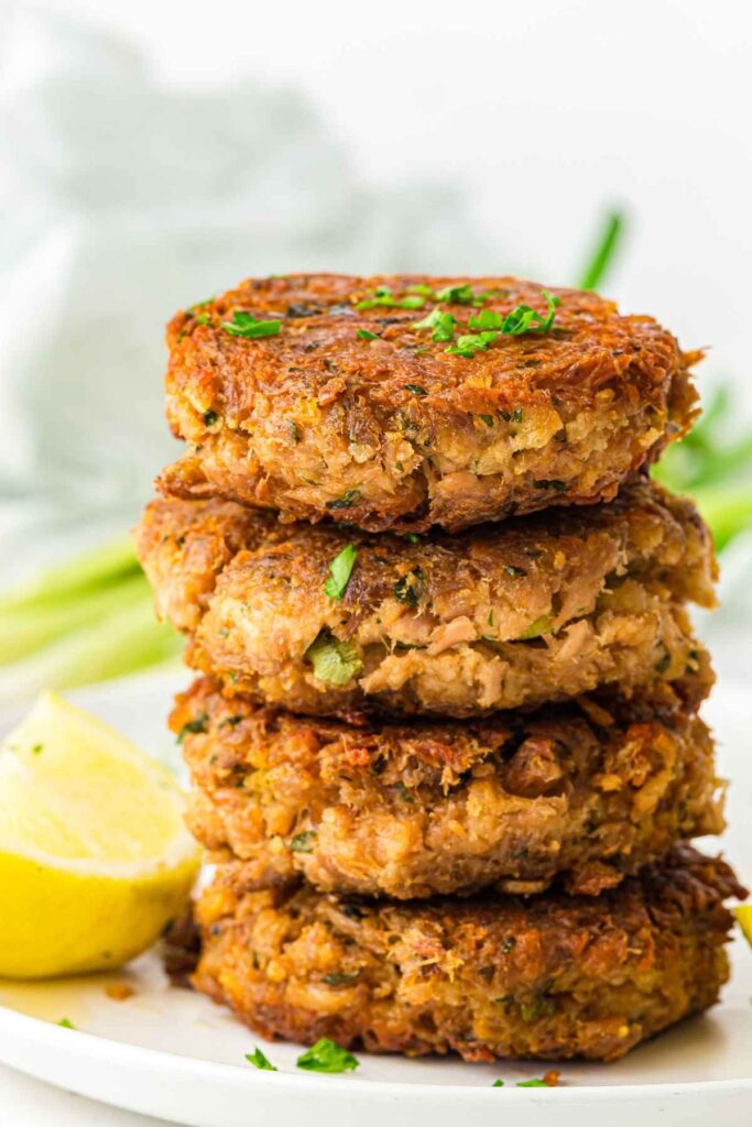 A stack of four golden-brown crab cakes garnished with chopped herbs, with a lemon wedge on a white plate.