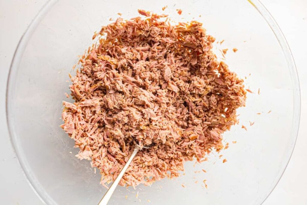 A glass bowl filled with shredded tuna and a metal fork resting inside.