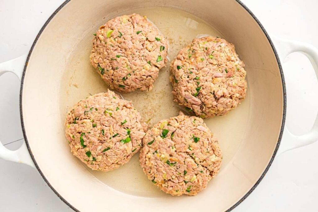 Four uncooked tuna patties with herbs are placed in a white skillet, ready to be cooked.