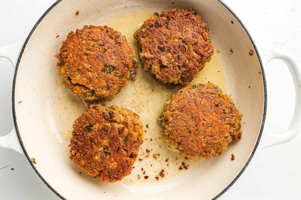 Four browned patties are cooking in a white skillet with a small amount of oil.