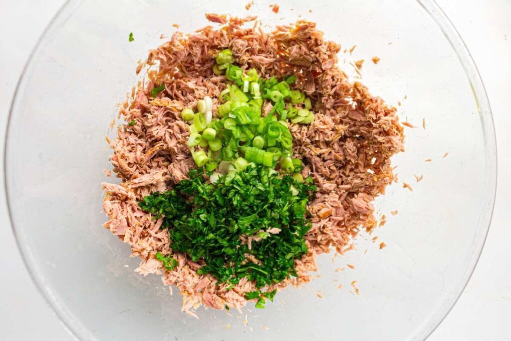 A glass bowl containing shredded tuna, chopped green onions, and chopped parsley, viewed from above.