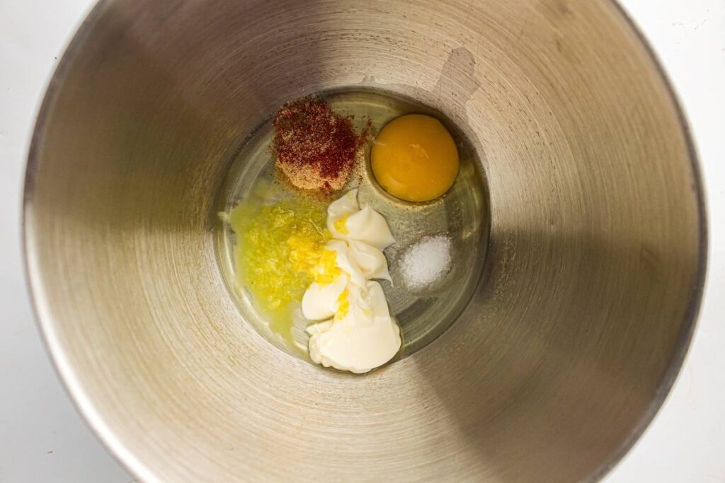 A metal mixing bowl containing an egg yolk, mayonnaise, minced garlic, lemon zest, seasoning, and salt.