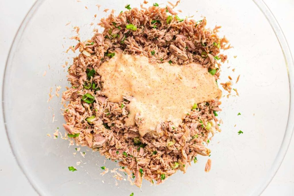 A glass bowl containing shredded tuna, chopped herbs, and a creamy sauce mixture.
