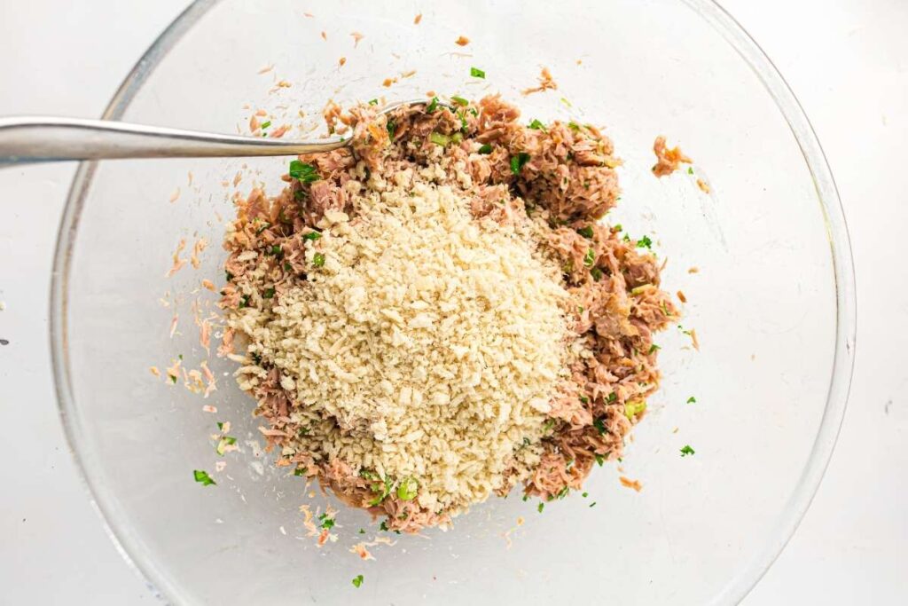 A glass bowl containing a mixture of tuna, chopped herbs, and a pile of breadcrumbs, with a metal spoon on the left side.